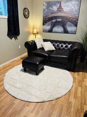 A living room with a black leather sofa, a white fluffy rug, a lamp, a clock, and a painting of the Eiffel Tower. - Private Suite w/Kitchen, Laundry, Gym, & Hot Tub at Tamarack (Edmonton)