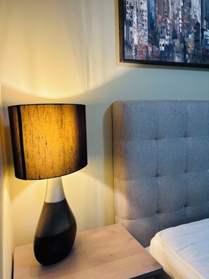 Un lit avec une tête de lit grise, une table de chevet avec une lampe et un tableau représentant un paysage urbain accroché au mur.