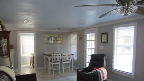 A living room with a ceiling fan, a dining area with a table and chairs, and large windows with blinds. - Beautiful vintage cottage remodeled in Old Ocean Springs (Ocean Springs)