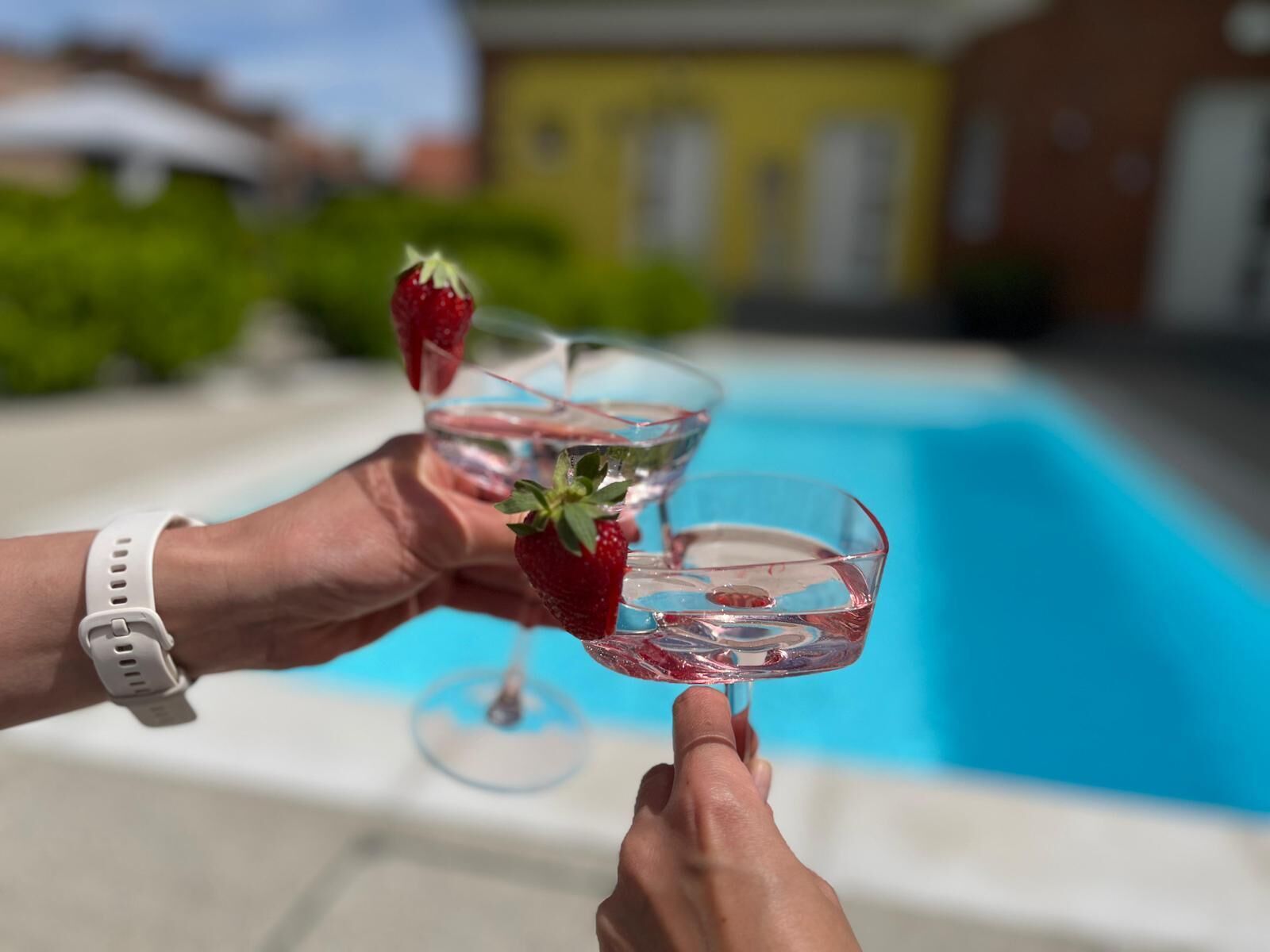 Two hands holding cocktail glasses with strawberries, near a swimming pool.