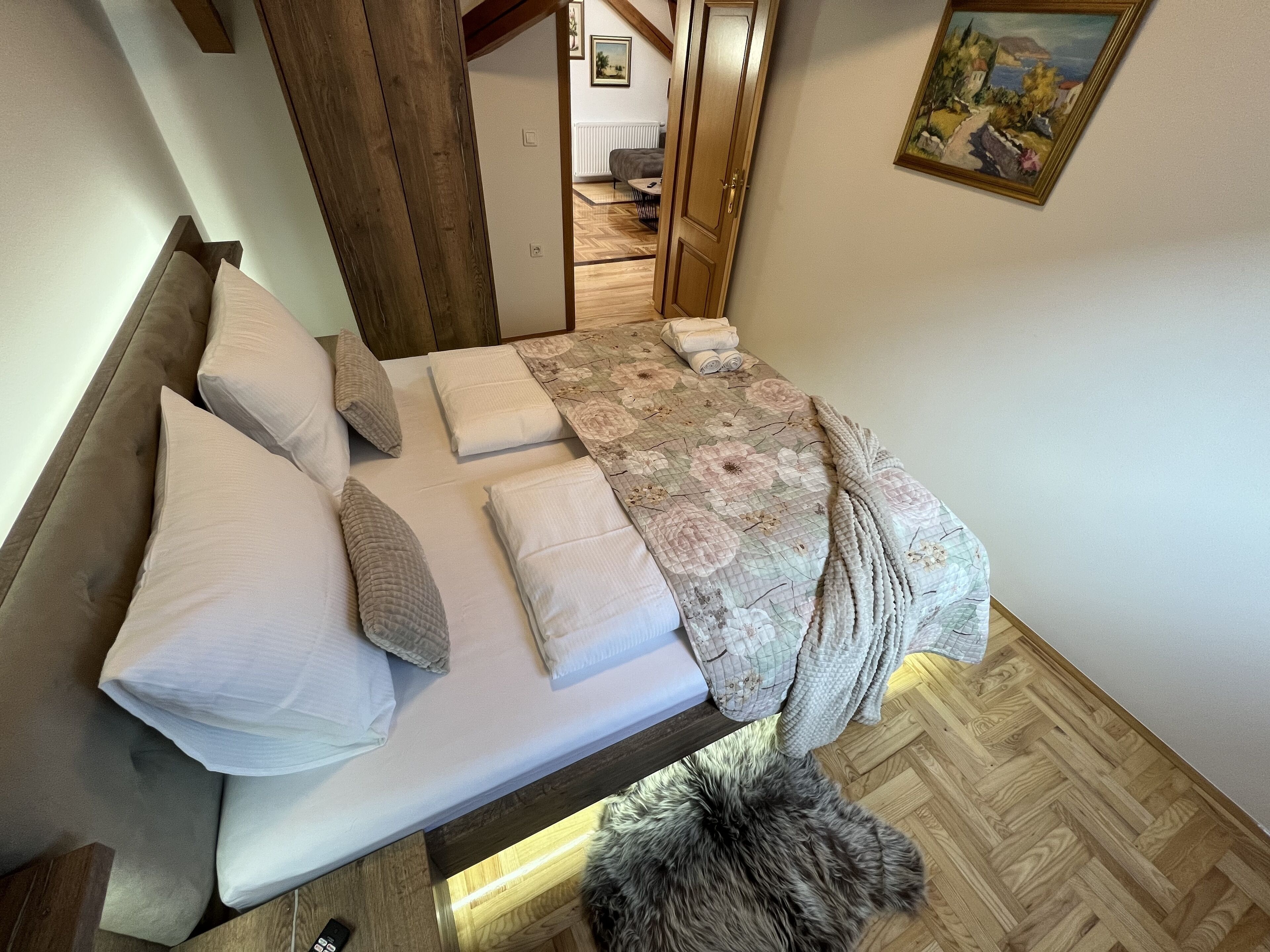 A neatly made bed with pillows and a blanket, a wooden headboard, and a fluffy rug on a wooden floor.