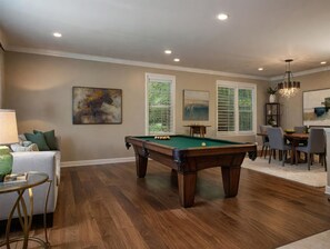 A pool table in a room with a dining area, two windows with blinds, and a chandelier.