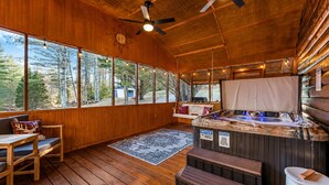 Interior de cabana com paredes de madeira, amplas janelas, uma banheira de hidromassagem e um deck de madeira.