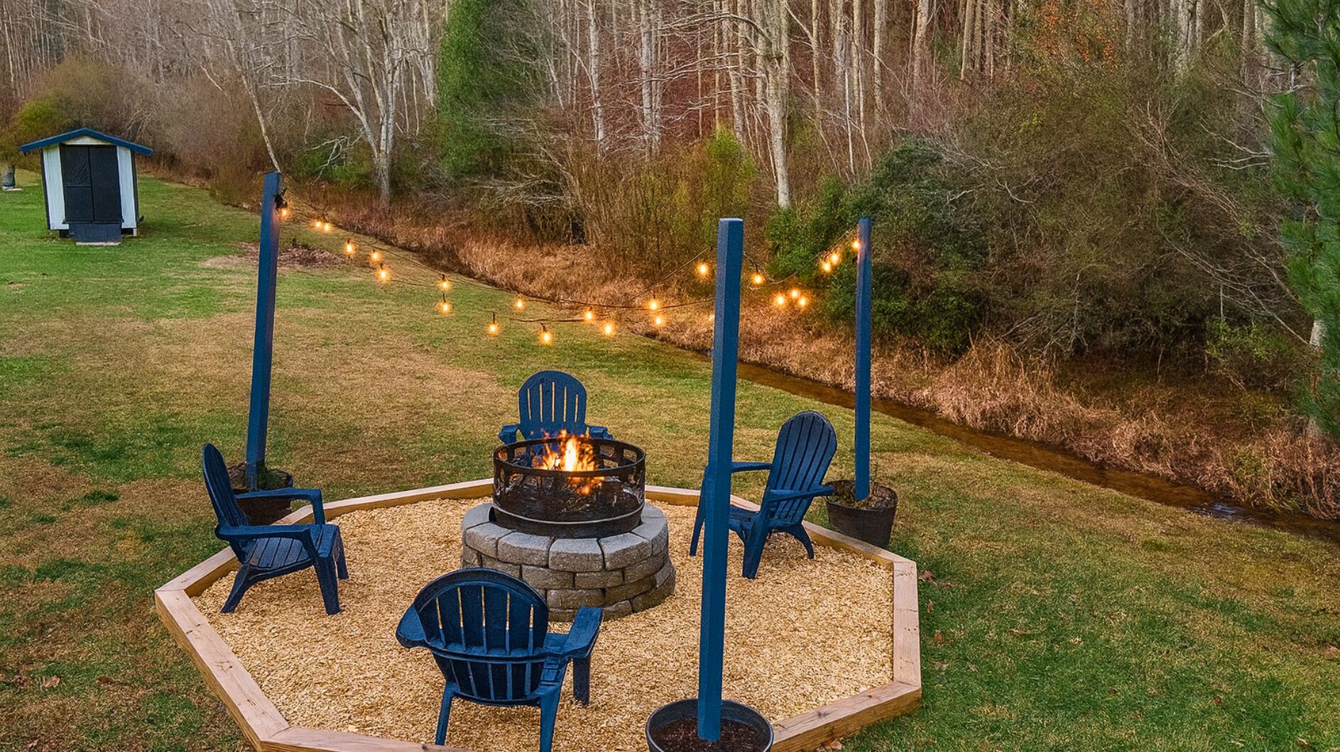 Un foyer autour duquel sont disposées quatre chaises Adirondack, le tout entouré d’une terrasse en bois et illuminé par une guirlande lumineuse.