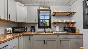 Uma cozinha com armários cinza claro, uma placa de fazenda sobre uma janela, e prateleiras de madeira com louças e uma abóbora.
