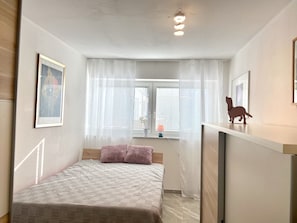 Une petite chambre bien éclairée, avec un lit, une fenêtre équipée de stores, une lampe de chevet et un tableau encadré au mur.
