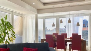 A modern living room with a sofa, dining table, and chairs. There are large windows with blinds, a potted plant, and pendant lights. - Ferienwohnung Riva - Wohlfühloase in Ruhiger Lage (Lindau)