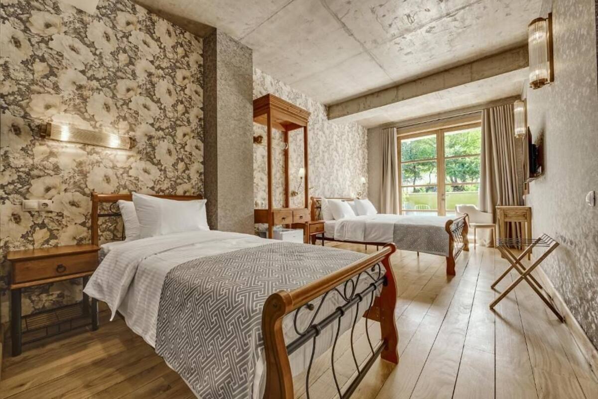 A room with two beds, a wooden headboard, a nightstand, a small table, a window with curtains, and a wall-mounted light fixture.