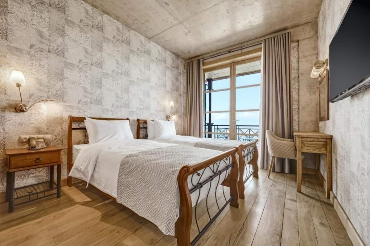 A bedroom with a large bed, wooden headboard, two bedside tables, a nightstand, a small wooden side table, a wall-mounted TV, and a large window with curtains.