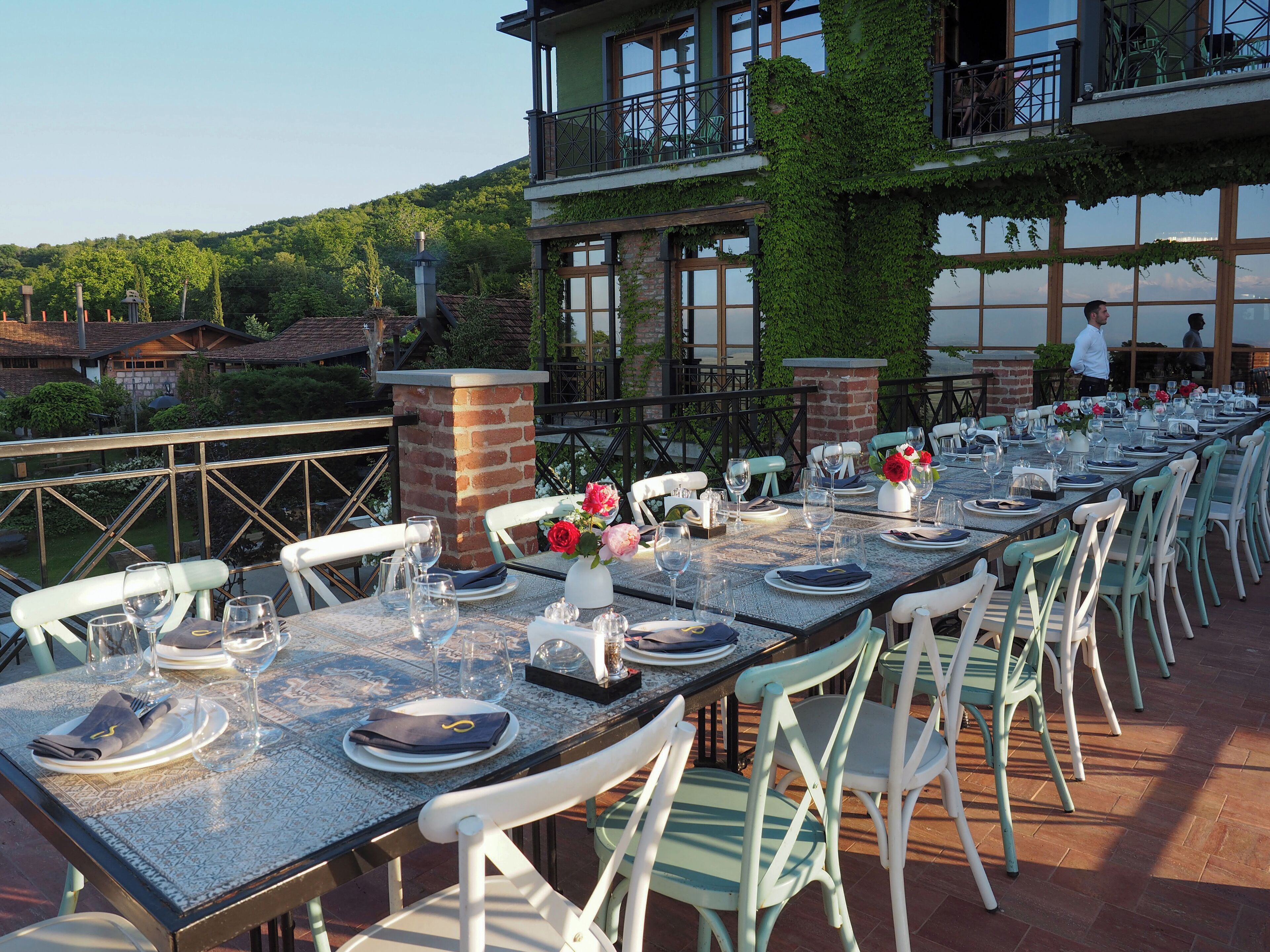 Una larga mesa de comedor al aire libre con sillas blancas, un pilar de ladrillo y vista a un edificio cubierto de hiedra.