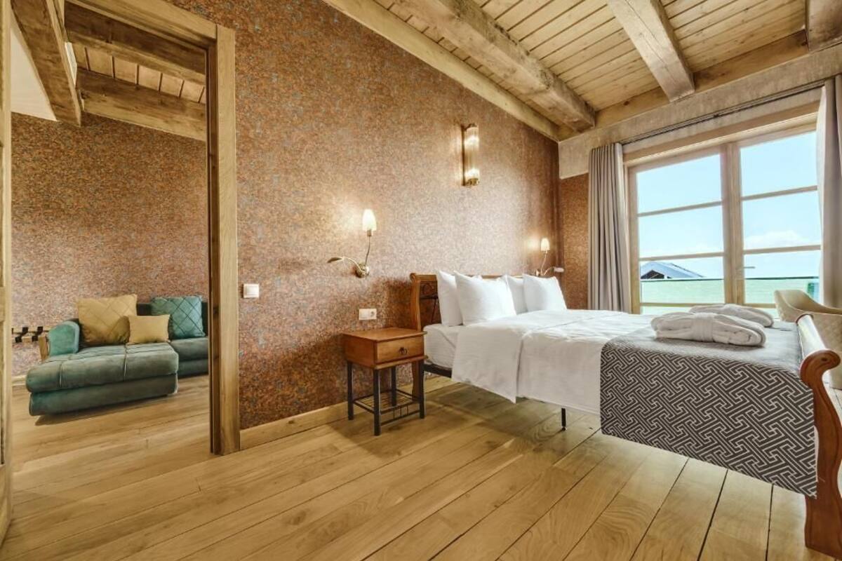 A hotel room with a wooden ceiling, a bed with a patterned bedspread, a small bedside table, a green sofa with cushions, and a view of the sea through large windows.