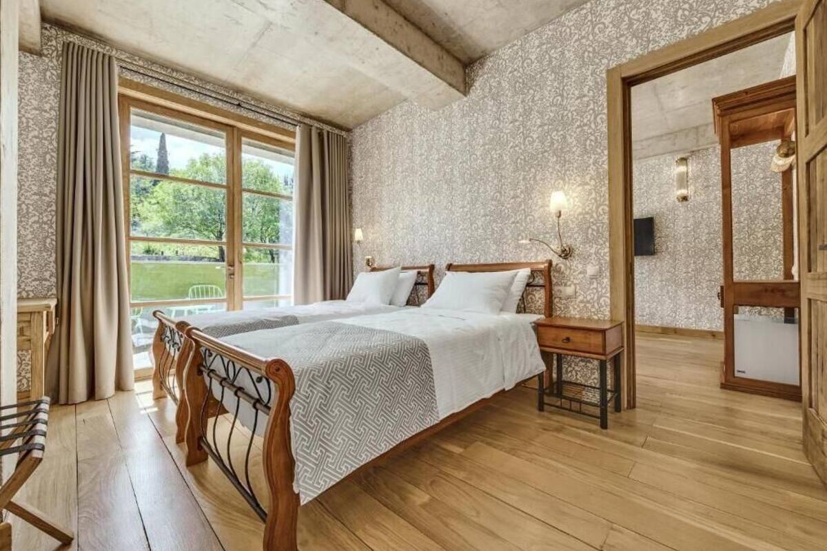 A double bed with a wooden frame, a bedside table, and a nightstand. There is a window with curtains, a mirror, and a view of greenery outside.