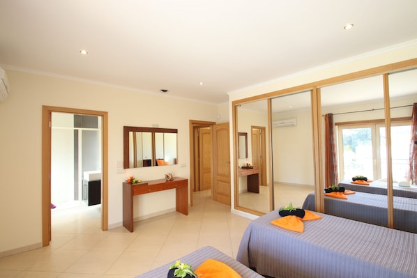A hotel room with a large mirror, a wooden desk, two beds with orange pillows, and a view of the outdoors.
