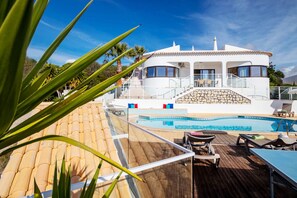 Zona junto a la piscina con terraza de madera, sillas de descanso y vista a una villa blanca con una escalera de piedra que lleva a la entrada.