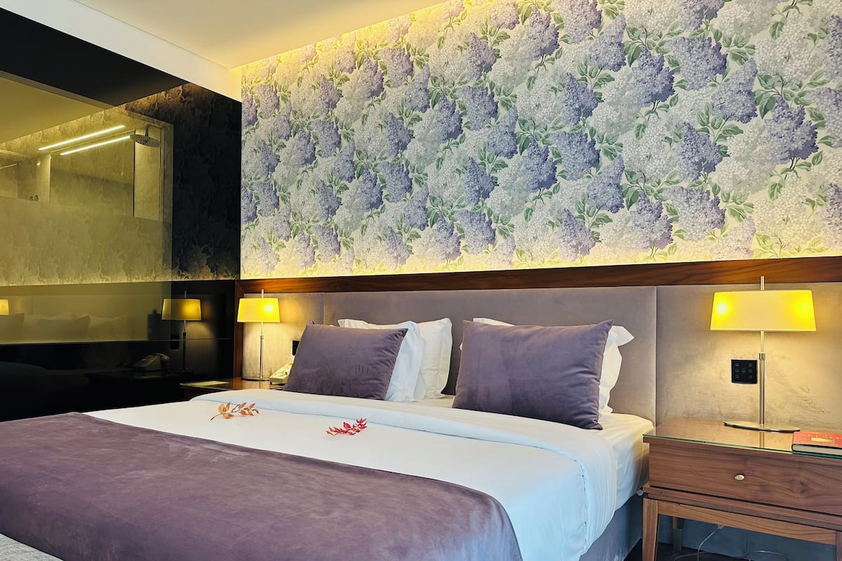 A hotel room with a large bed, two bedside lamps, a wooden nightstand, and a wall with floral wallpaper.