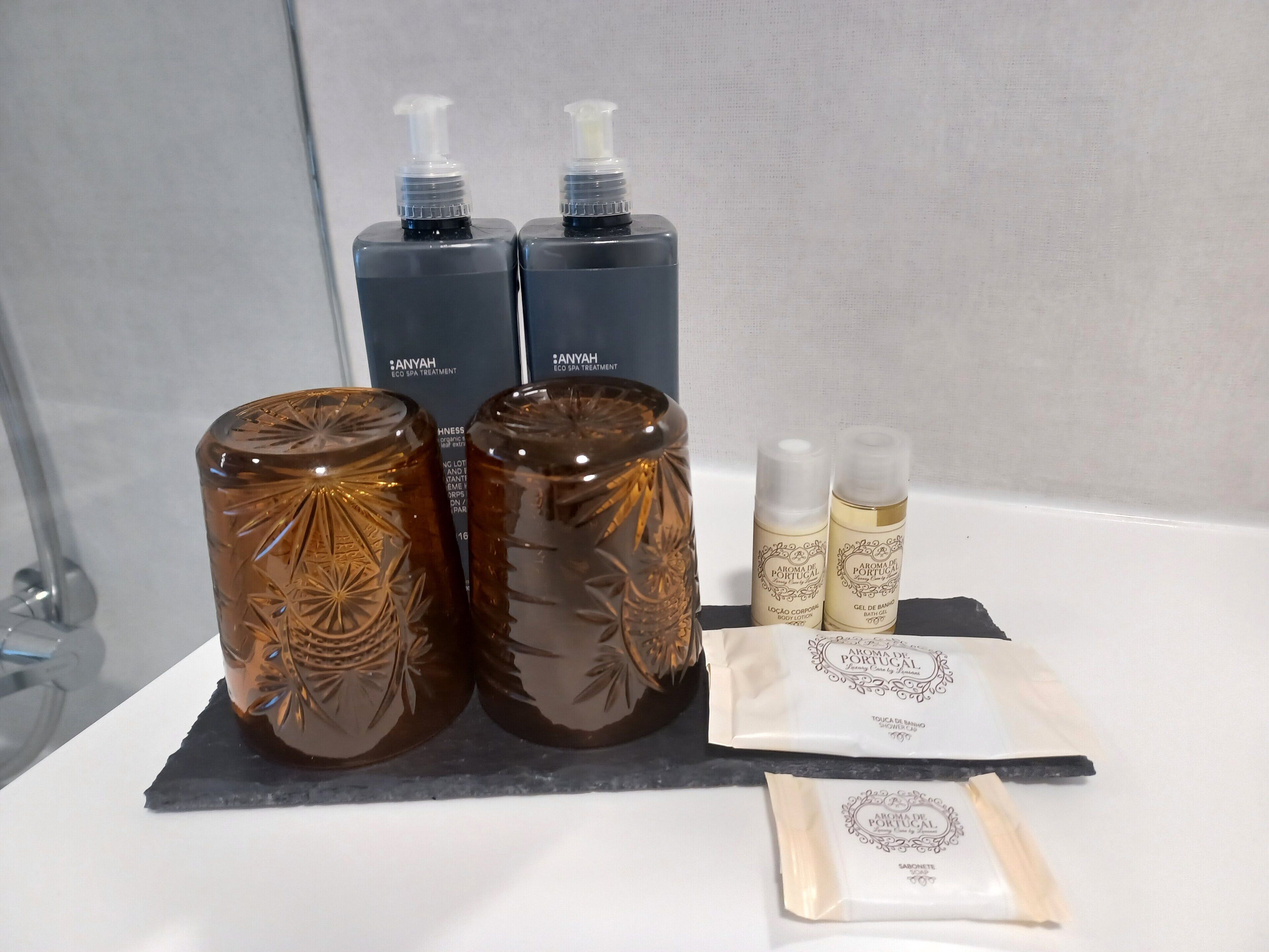 Un baño con un lavamanos blanco, dos recipientes de vidrio marrón, dos botellas de loción corporal y una toalla blanca con un logotipo.