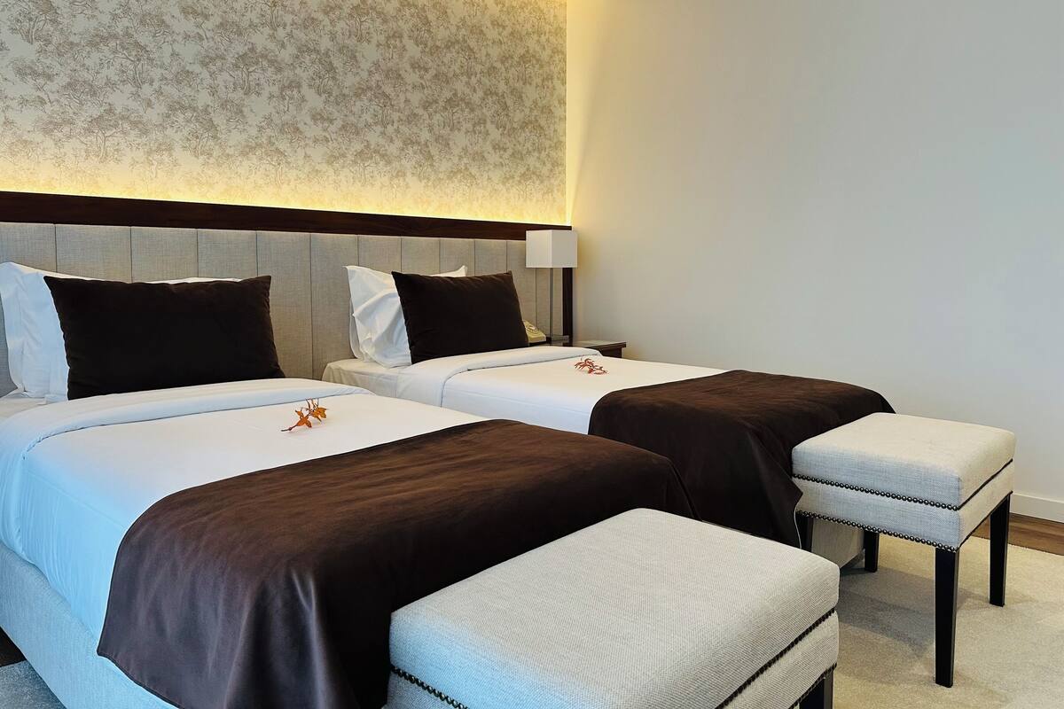 A hotel room with two beds, a headboard, bedside tables, and a small bench.