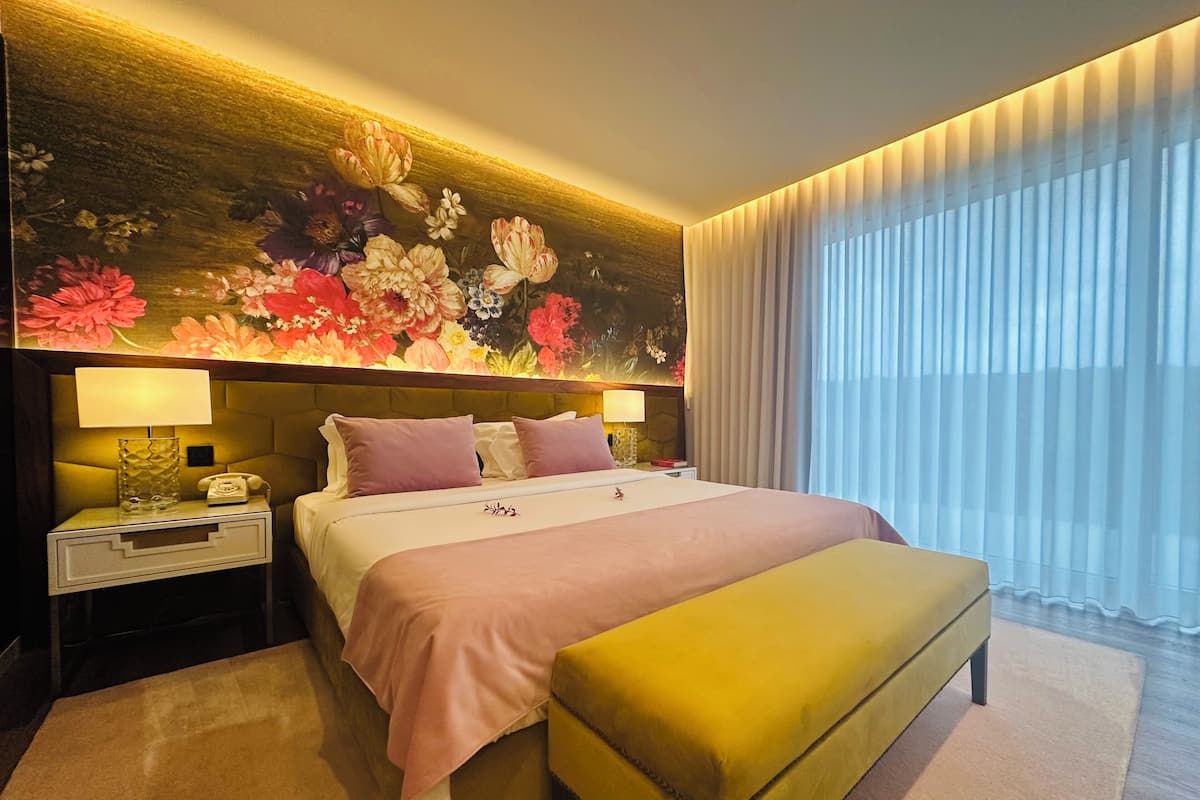 A hotel room with a bed, bedside lamps, a nightstand, a bench, a floral mural, and a large window with curtains.