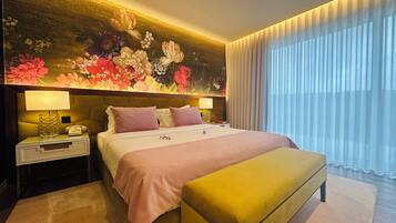 Quarto de hotel com cama, luminárias laterais, mesa de cabeceira, um banco, um mural floral e uma janela grande com cortinas.
