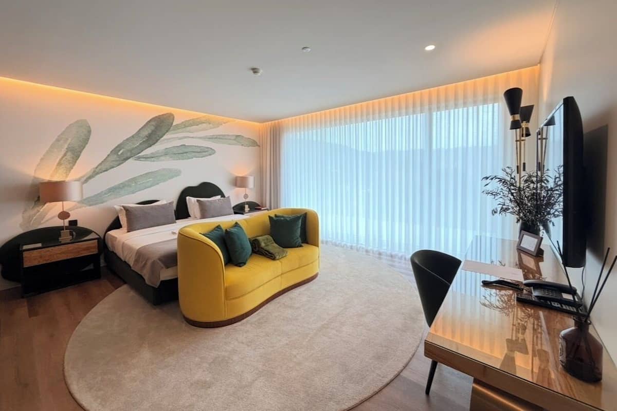 A modern hotel room with a bed, a yellow sofa, a desk, and a large window with blinds.