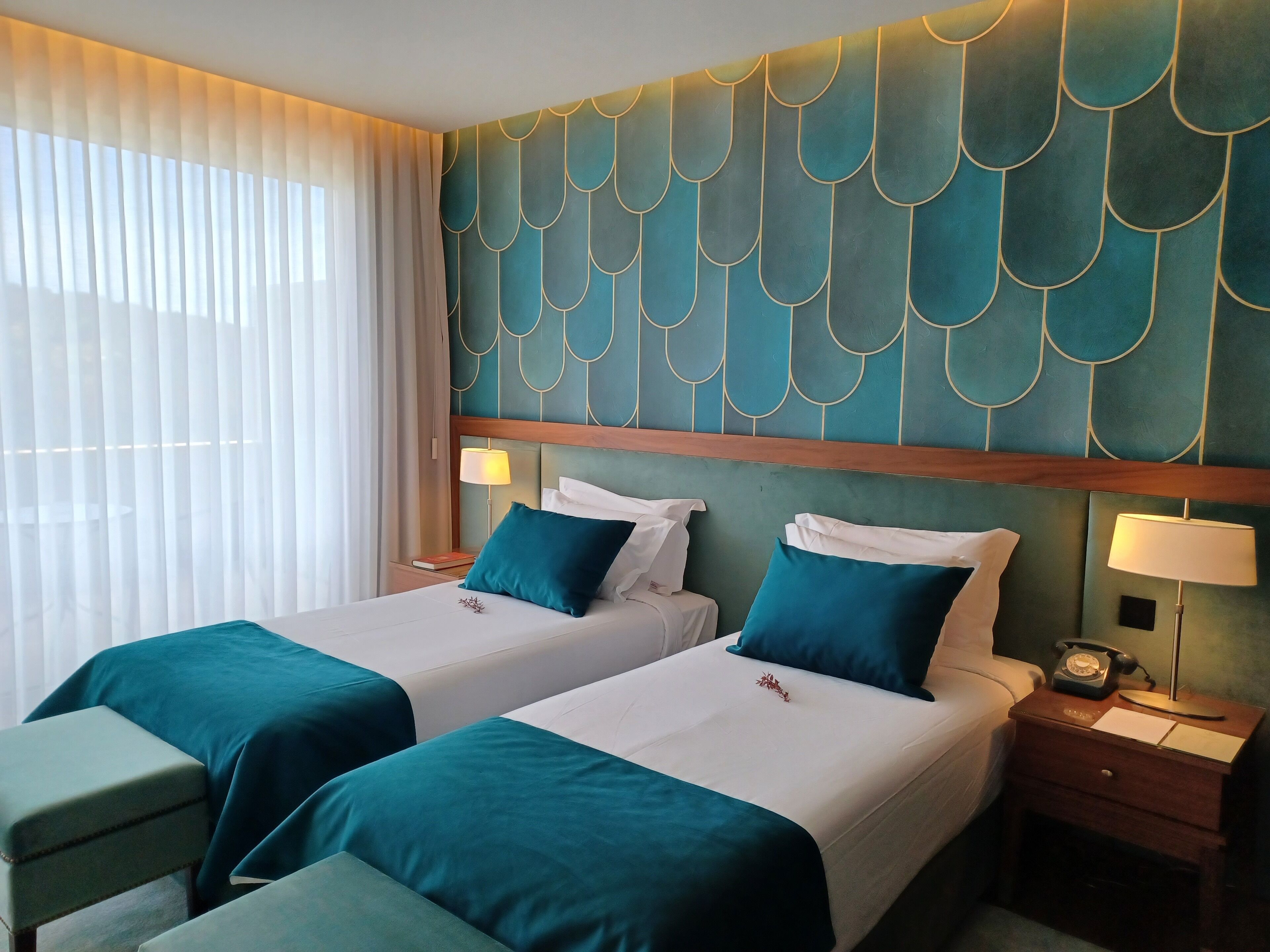 A hotel room with two beds, teal bedding, a decorative teal wall, and a bedside table with a lamp.