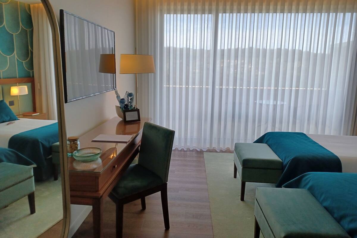 A hotel room with a bed, a desk with a lamp, a chair, a small table with a vase, and a large window with curtains.