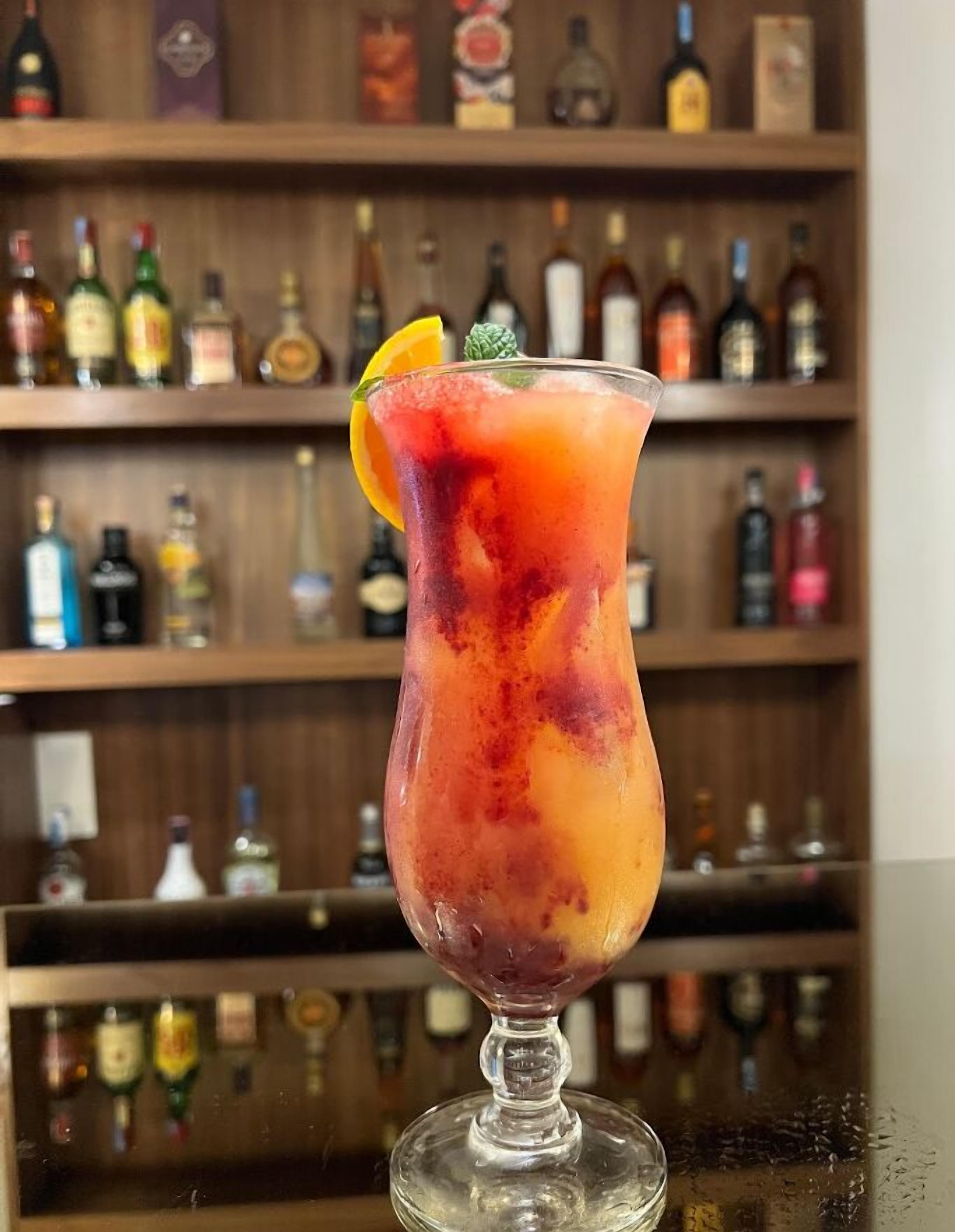 A cocktail in a glass with a fruit garnish, set against a backdrop of a bar with shelves of liquor bottles.