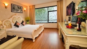 Une chambre à coucher avec un grand lit, une table de chevet avec une lampe, une commode, une table en verre avec une bouteille, et une fenêtre ornée de motifs en fer forgé.