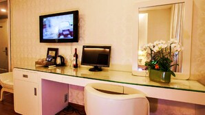 Une chambre d’hôtel avec un bureau en verre, deux écrans d’ordinateur, un petit réfrigérateur, une chaise blanche et un miroir agrémenté d’un arrangement floral.