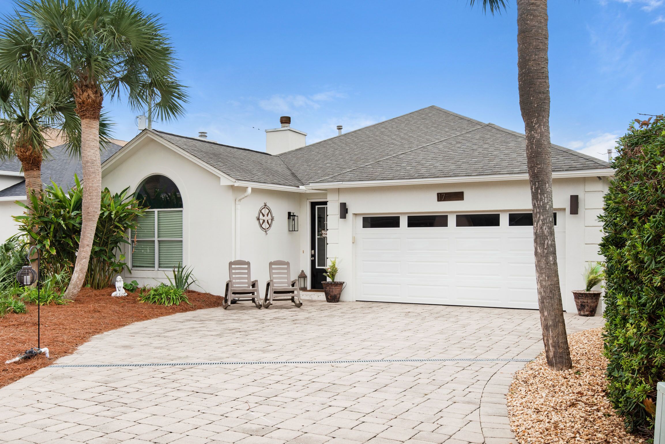 A single-story house with a garage, a front porch with two chairs, and a palm tree.