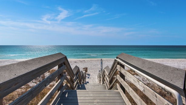 Une promenade en bois qui mène à une plage où l’eau est d’un bleu limpide et le rivage est sablonneux.