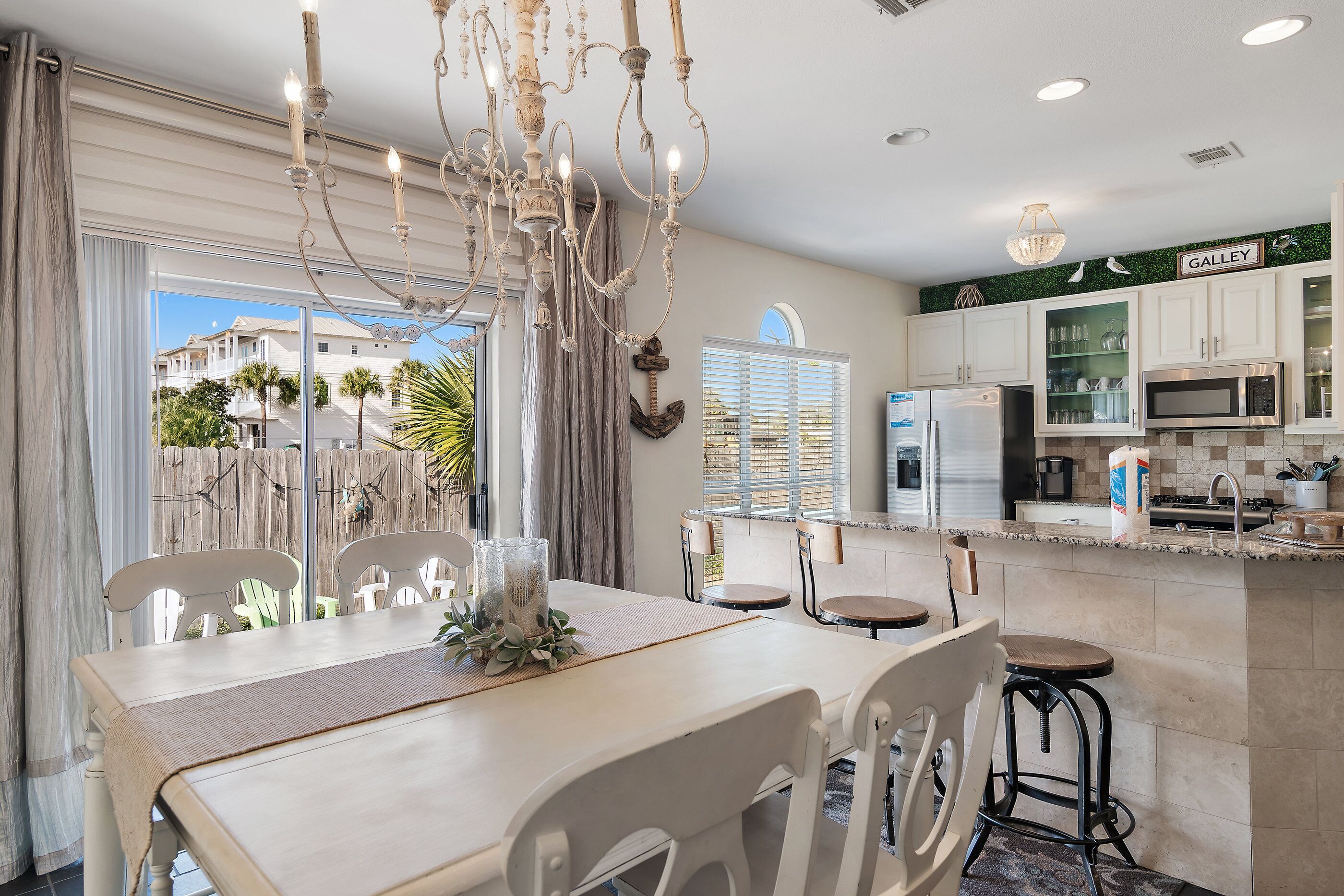 A modern kitchen with white cabinets, a microwave, and a refrigerator. There is a dining area with a table and chairs, and a chandelier above.