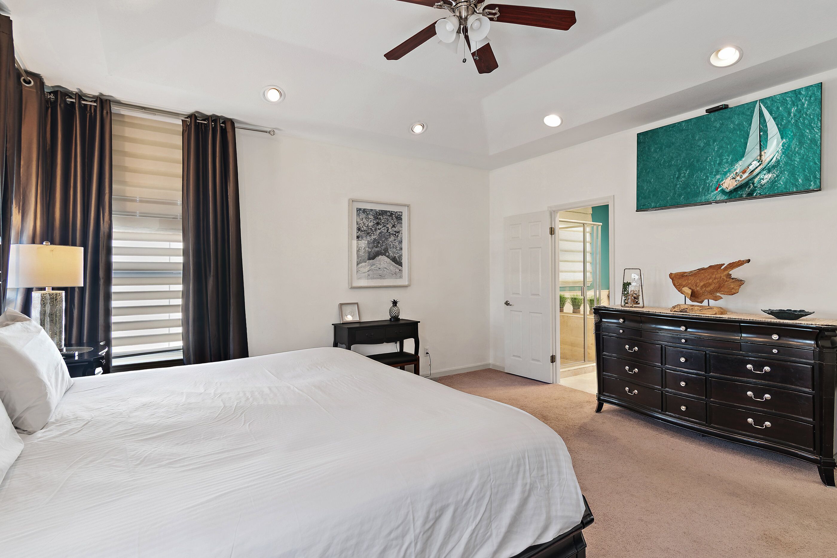 A bedroom with a large bed, a dresser, a ceiling fan, a painting, and a door leading to another room.