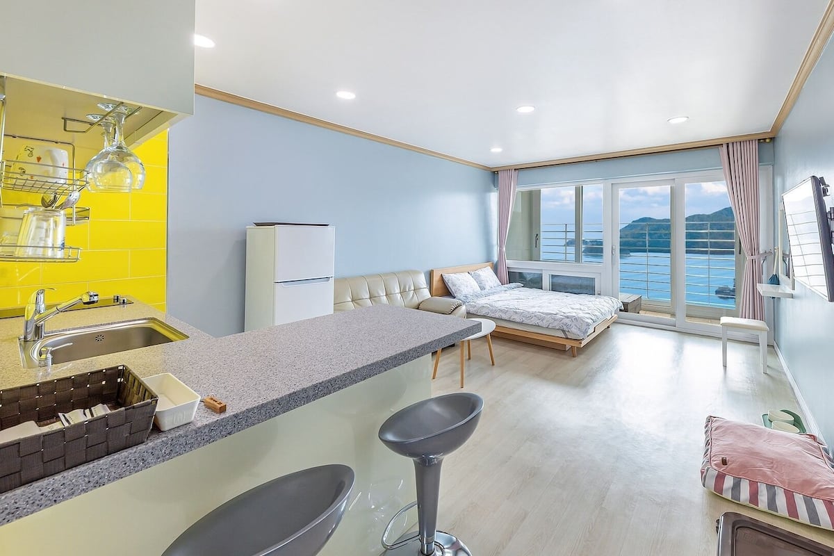 A compact room with a kitchenette, a bed, a bar stool, and a view of the sea.