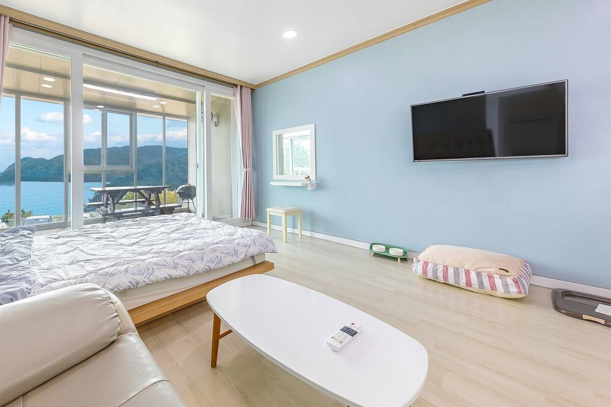 A bedroom with a bed, a TV, a table, a sofa, and a view of the outdoors.