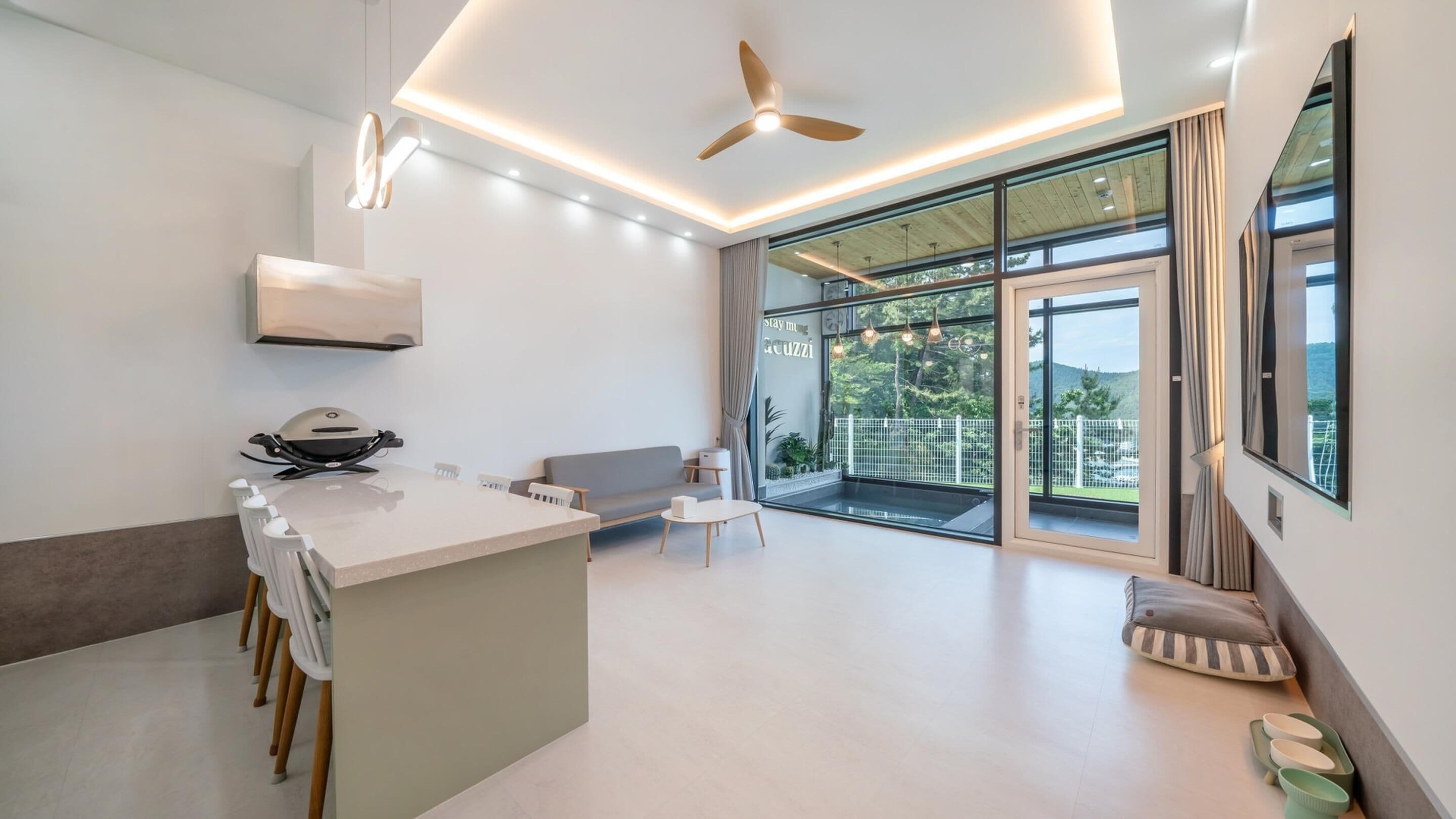 A modern kitchen with a central island, a ceiling fan, and large windows offering a view of greenery.