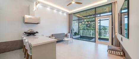 A modern kitchen with a central island, a ceiling fan, and large windows offering a view of greenery.