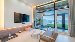 A modern living room with a large TV, a sofa, a small table, and a balcony with a view of the sea. - Yeosu Seuteimeong (Yeosu)