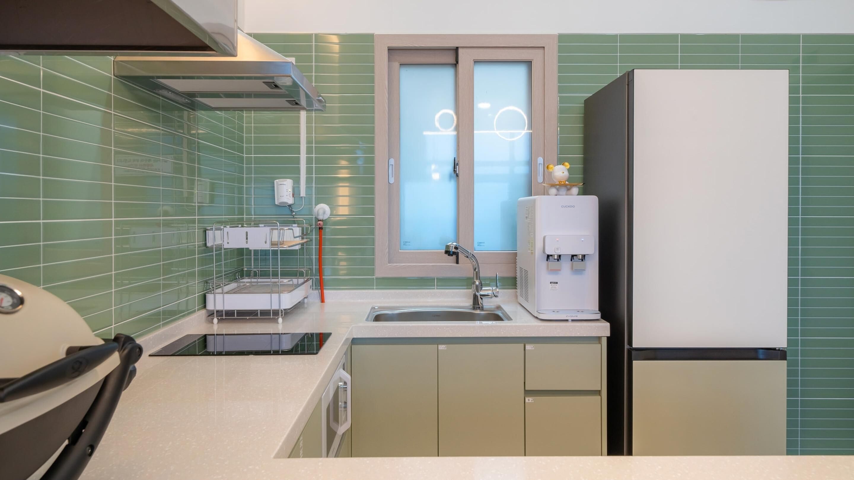 Una cocina compacta con paredes de azulejos verdes, un lavamanos empotrado, dispensador de agua y refrigerador.