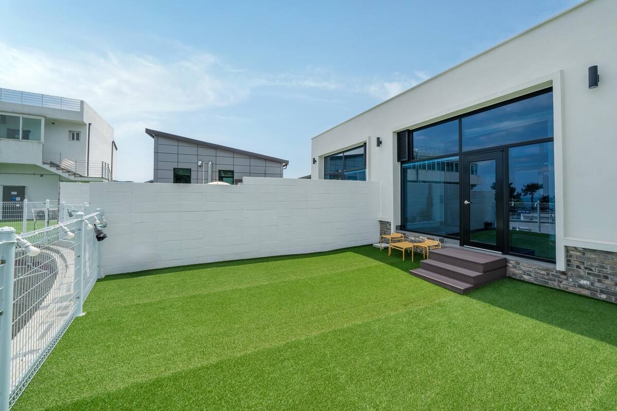 A modern house with a white exterior, large glass windows, and a small patio with artificial grass and outdoor furniture.
