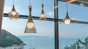 A view through a window with three hanging pendant lights, a metal railing, and a scenic view of a body of water and distant mountains. - Yeosu Seuteimeong (Yeosu)