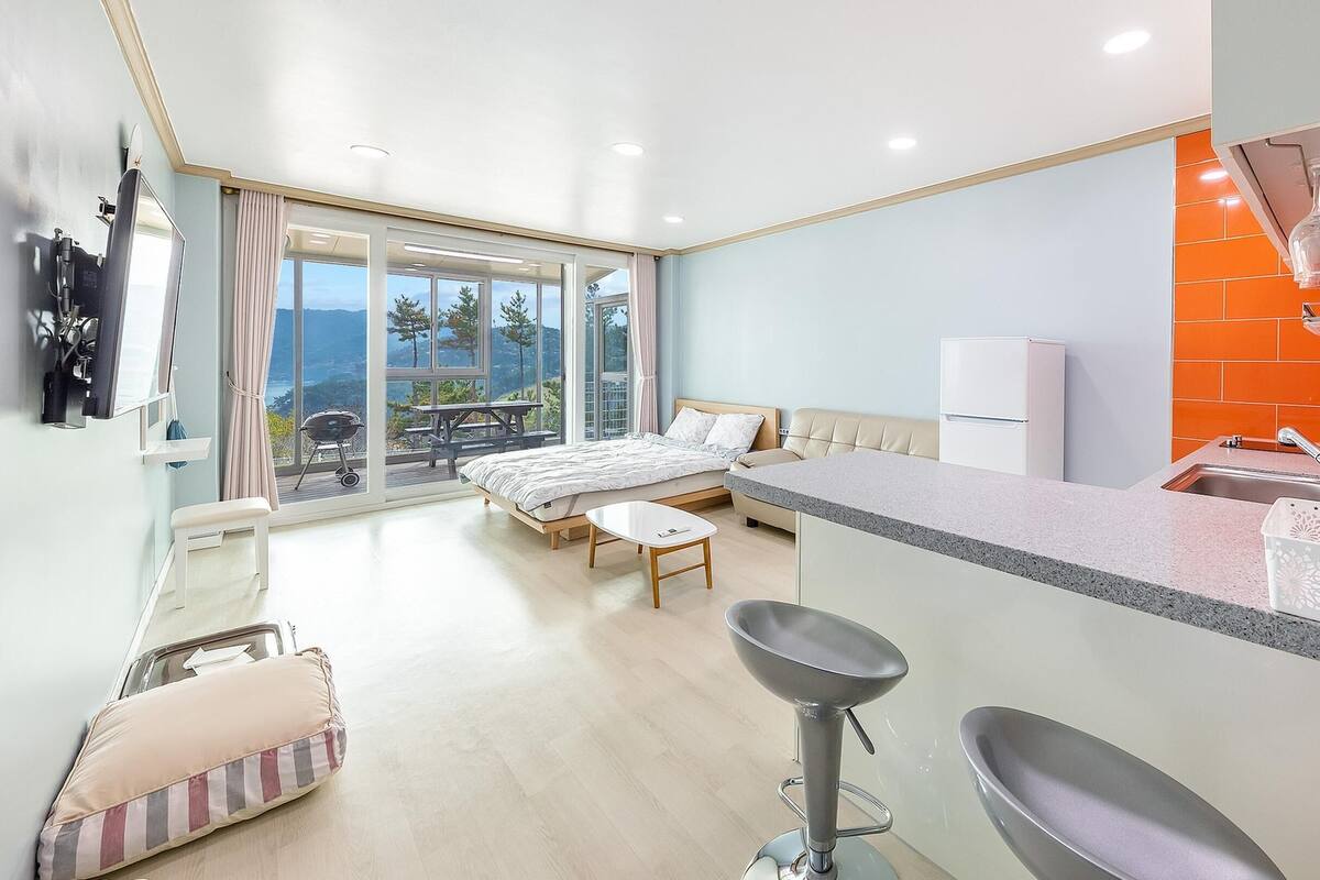 A modern hotel room with a bed, a small table, a kitchenette, and a balcony with a view.