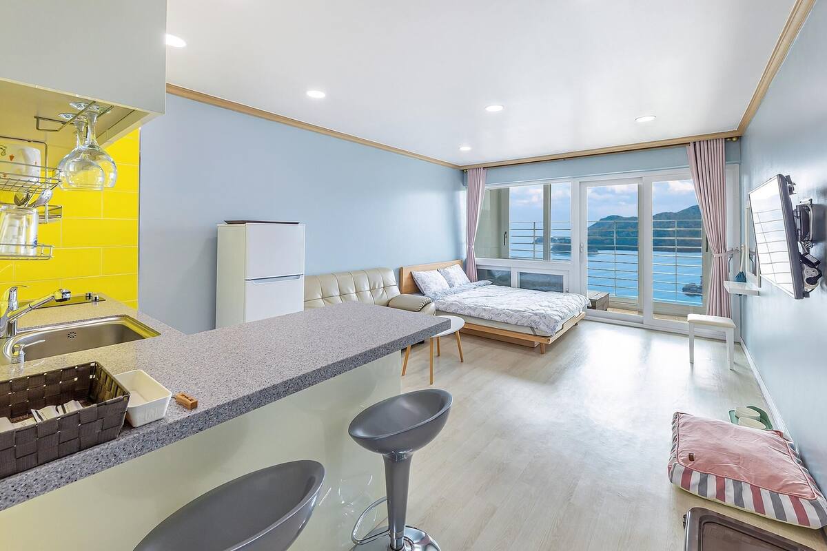 A compact room with a kitchenette, a bed, and a large window offering a view of the sea.