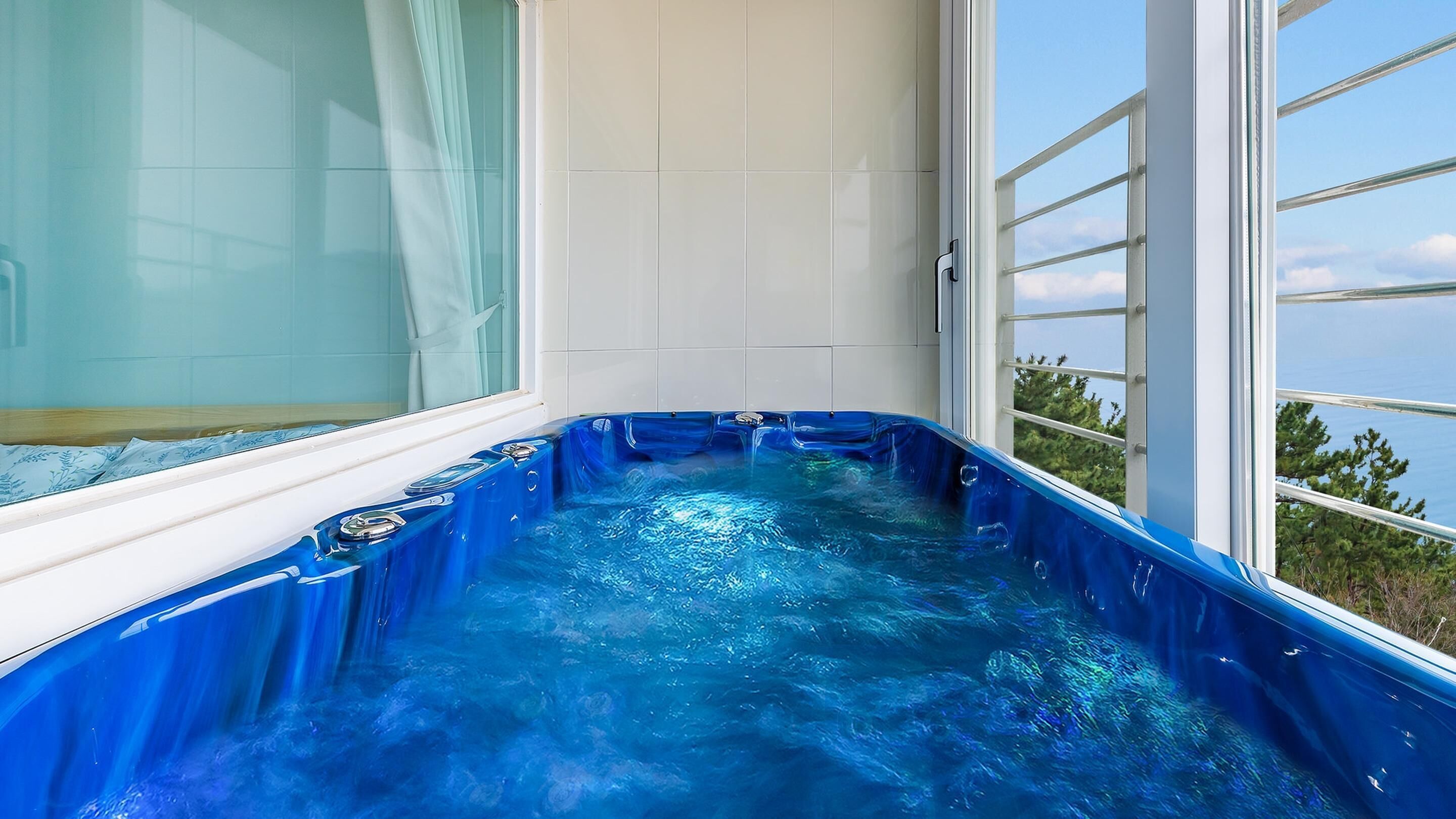 A hot tub with a blue interior, adjacent to a window with a view of trees and a clear sky.