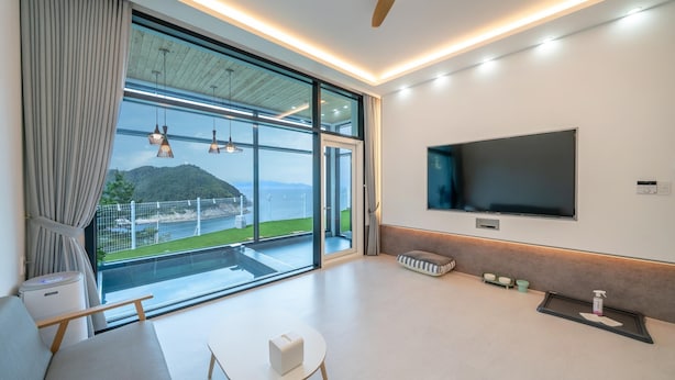 A modern living room with a large TV, a sliding glass door to a balcony, and a view of a mountainous landscape. - Yeosu Seuteimeong (Yeosu)