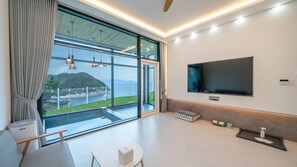 A modern living room with a large TV, a sliding glass door to a balcony, and a view of a mountainous landscape. - Yeosu Seuteimeong (Yeosu)