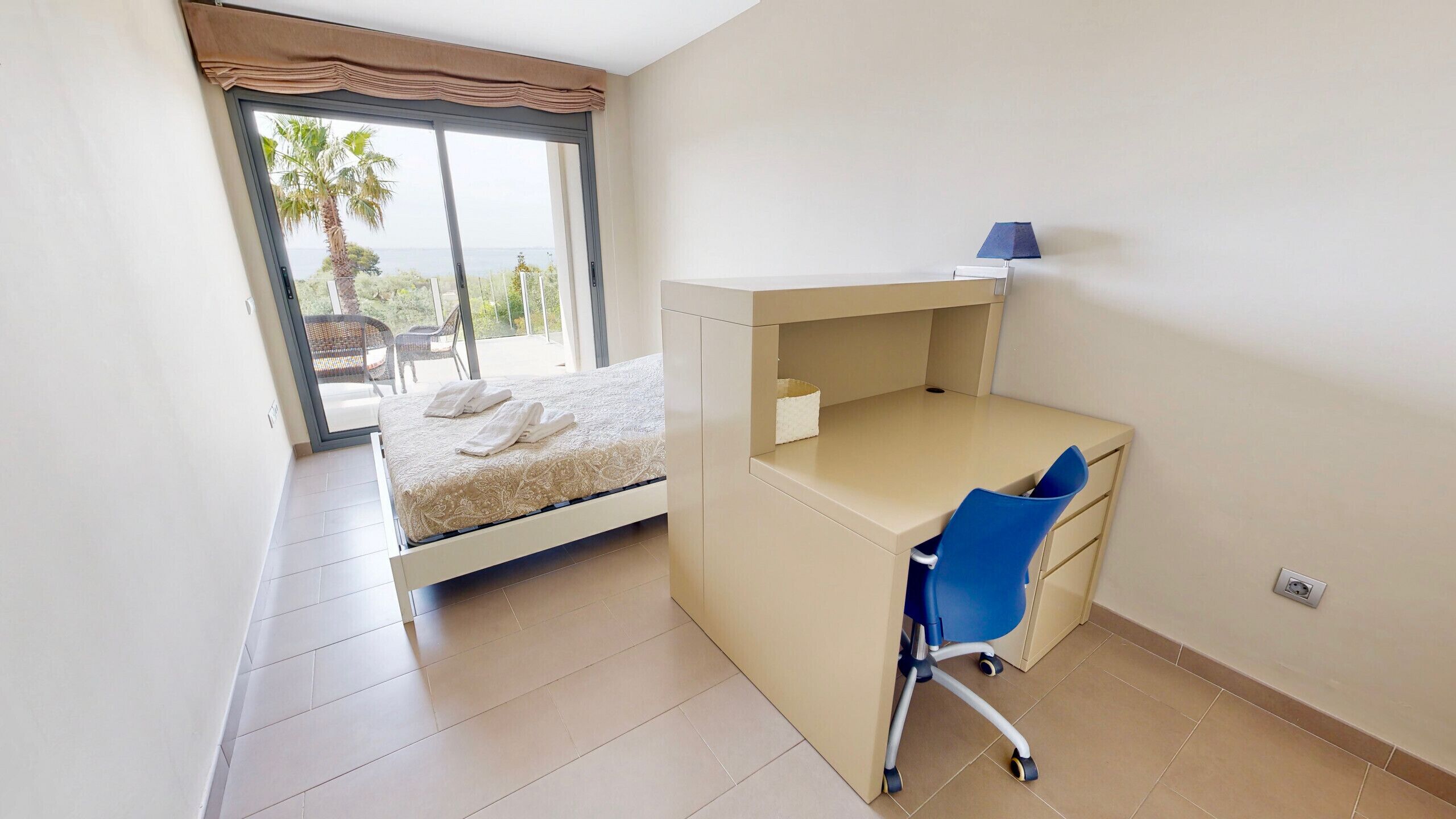 Un dormitorio con cama, escritorio y silla. Hay una ventana con vistas a palmeras y un balcón.