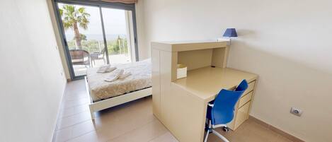 Un dormitorio con cama, escritorio y silla. Hay una ventana con vistas a palmeras y un balcón.