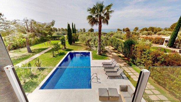 A rectangular swimming pool with a clear blue water surface, surrounded by a paved deck. There are several lounge chairs and a small sofa arranged around the pool. The pool is set in a landscaped garden with various trees and shrubs. - VILLA NIRVANA (El Perelló)
