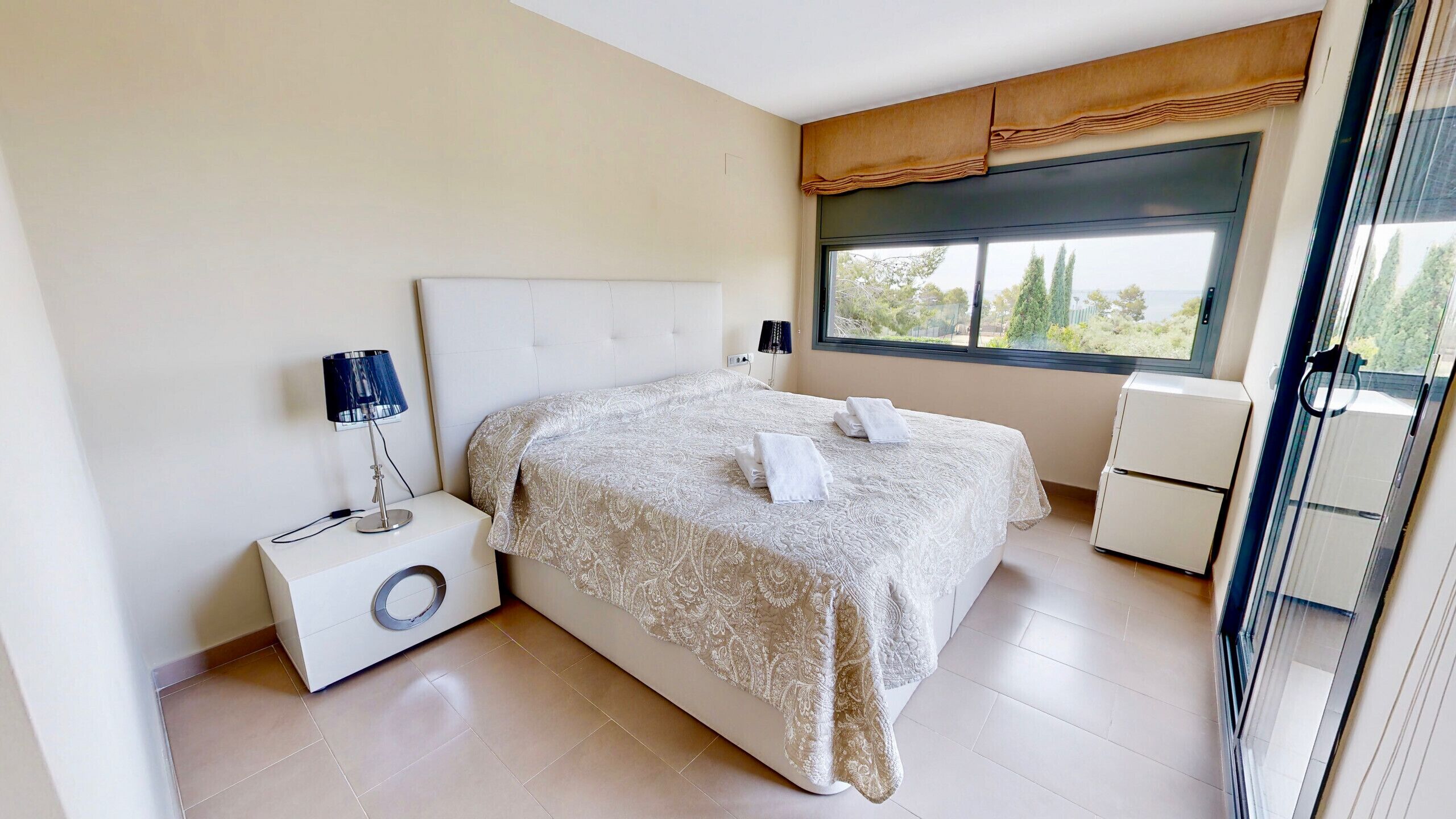 A modern bedroom with a large bed, a bedside table with a lamp, a small white cabinet, and a window with a view of trees.